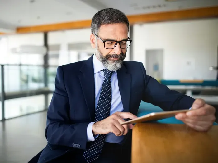 Confident middle aged businessman in a suit using a tablet in a bright office lounge, reviewing local services